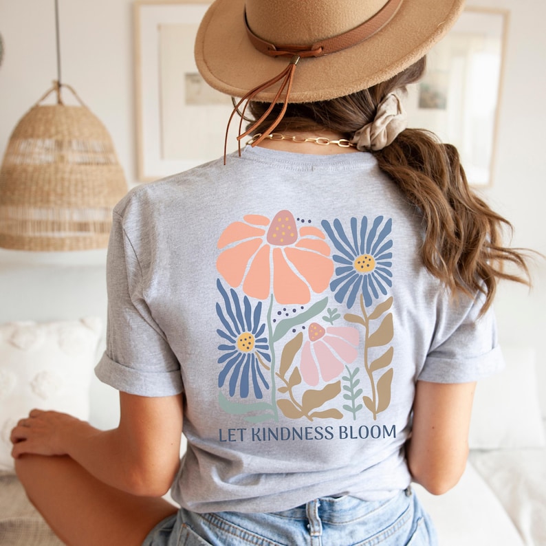 May include: A grey t-shirt with a floral design on the back. The design includes large orange and pink flowers, blue flowers, and the text "LET KINDNESS BLOOM". The shirt has rolled-up sleeves.