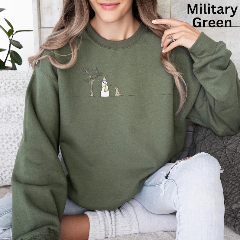 May include: A military green crewneck sweatshirt featuring a minimalist line art graphic of a bare tree, a snowman wearing a yellow scarf and hat, and a small dog standing on a horizontal line. The text Military Green appears in the top corner.