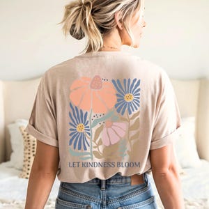 May include: Beige t-shirt with a floral design in peach, blue, and pink. The graphic features stylized flowers and the text "LET KINDNESS BLOOM" in a simple font. The shirt has rolled-up sleeves.