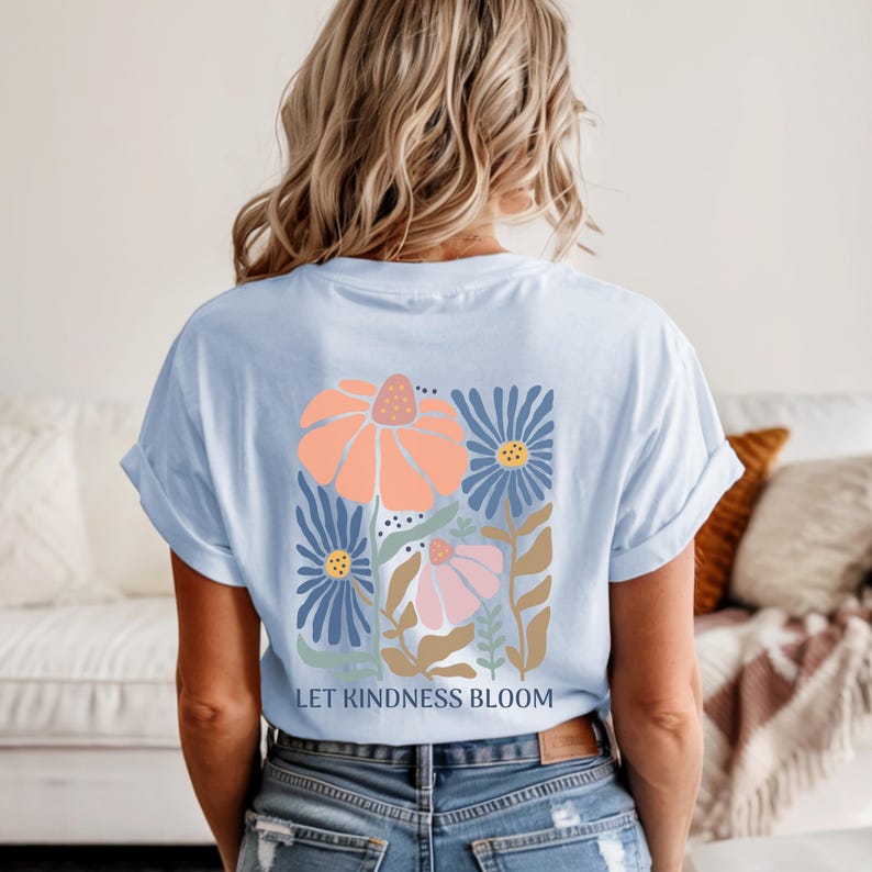 May include: Pale blue t-shirt featuring a floral design in peach, blue, and pink. The graphic includes the text "LET KINDNESS BLOOM". The shirt is a casual style.