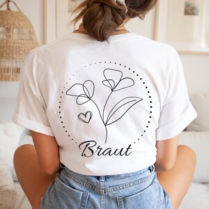 May include: White t-shirt with a black floral design and the word "Braut". The design includes two flowers, a heart, and a dotted circle. The shirt is made of a soft material.