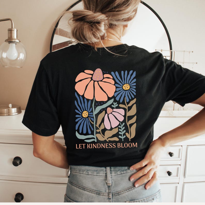 May include: Black t-shirt featuring a floral design in peach, blue, and pink, with the text "LET KINDNESS BLOOM". The graphic shows stylized flowers and foliage. The shirt is a casual wear item.