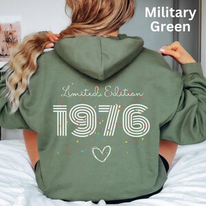 Könnte beinhalten: Militärgrüner Kapuzenpullover mit dem weißen Text "Limited Edition 1976", bunten Konfetti und einer Herzgrafik. Der Kapuzenpullover hat eine Kapuze mit Kordelzug und lange Ärmel. Der Text "Military Green" befindet sich oben rechts.