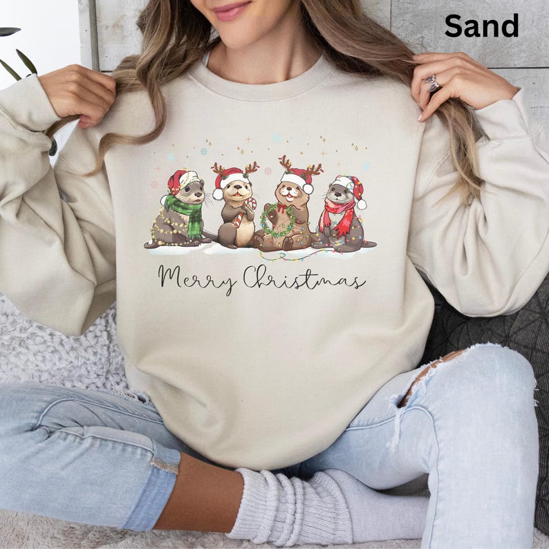 Pu&ograve; includere: Felpa color sabbia con un design natalizio. La grafica raffigura quattro lontre con cappelli di Babbo Natale, corna di renna e sciarpe, con la scritta "Merry Christmas" sotto. Le lontre sono circondate da luci decorative, creando un'atmosfera festosa.
