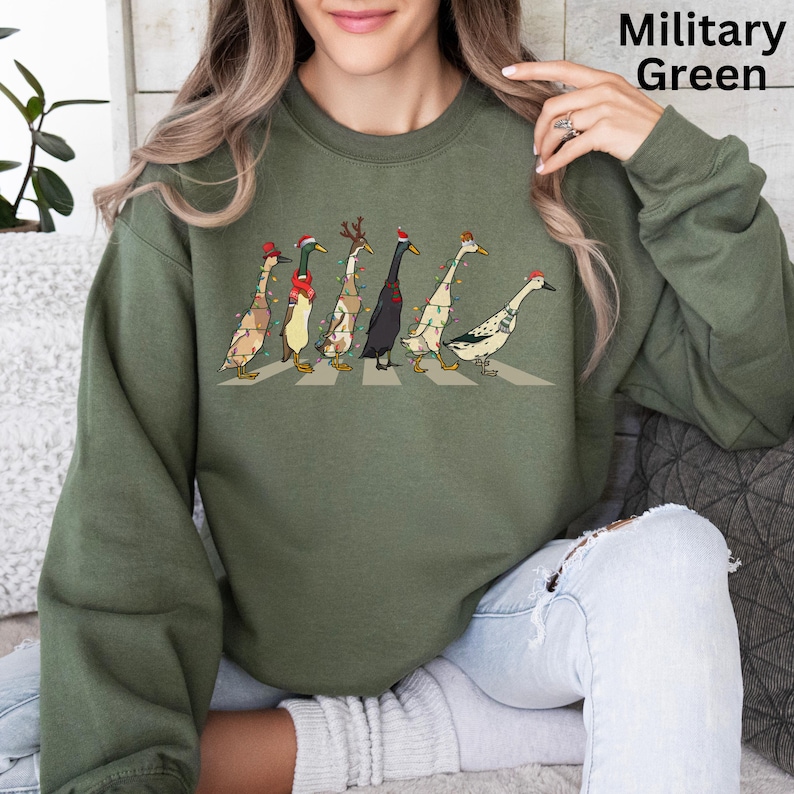 Peut inclure: Un sweat-shirt vert avec un motif de No&euml;l repr&eacute;sentant une file de canards portant des bonnets de P&egrave;re No&euml;l et des lumi&egrave;res de No&euml;l traversant une rue. Le texte "Military Green" est visible dans le coin sup&eacute;rieur droit de l'image.