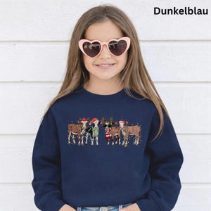 Puede incluir: Una sudadera azul marino con un dise&ntilde;o festivo de vacas adornadas con luces navide&ntilde;as, gorros de Pap&aacute; Noel y cuernos de reno. El texto "Dunkelblau" es visible en la parte superior. La persona de la imagen lleva gafas de sol en forma de coraz&oacute;n.