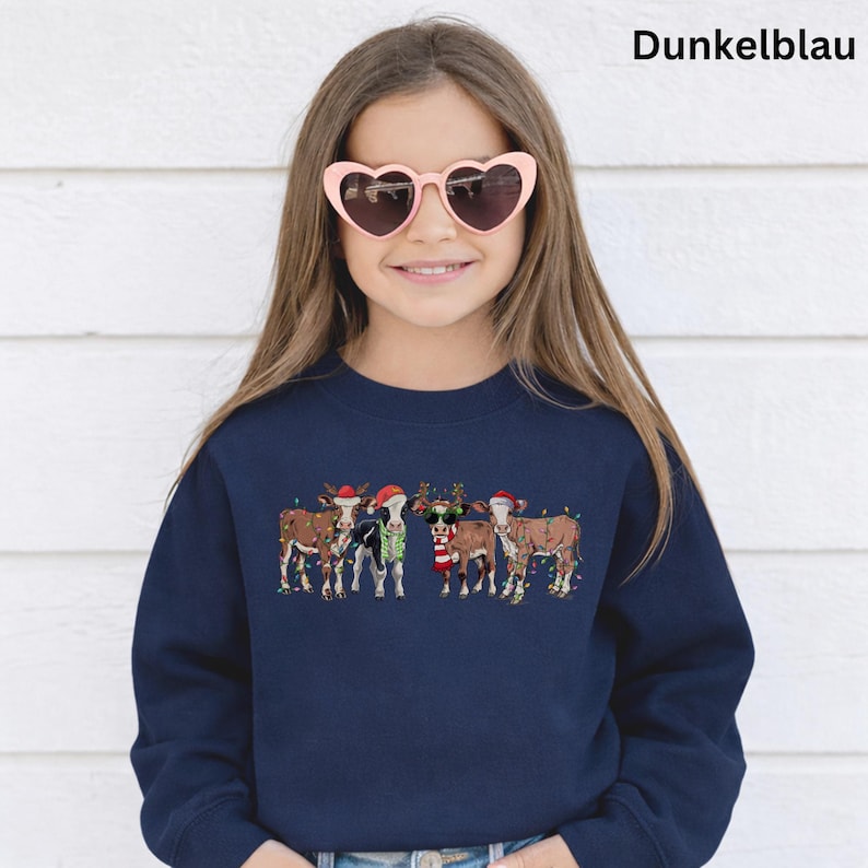 Puede incluir: Una sudadera azul marino con un dise&ntilde;o festivo de vacas adornadas con luces navide&ntilde;as, gorros de Pap&aacute; Noel y cuernos de reno. El texto "Dunkelblau" es visible en la parte superior. La persona de la imagen lleva gafas de sol en forma de coraz&oacute;n.