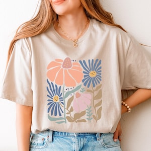 May include: Beige t-shirt featuring a floral design with peach, blue, and pink flowers. The graphic includes stylized leaves and stems. The shirt has short sleeves and a relaxed fit, suitable for casual wear.