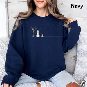 May include: A navy blue crewneck sweatshirt featuring a minimalist graphic print of a bare tree, a snowman wearing a hat and scarf, and a small dog standing on a thin horizontal line, with the word Navy printed in the upper right corner.