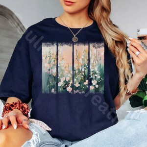 May include: Navy blue t-shirt featuring a floral design. The artwork is divided into four vertical panels with a pastel color palette of pink, green, and white flowers. The shirt has short sleeves and a relaxed fit.