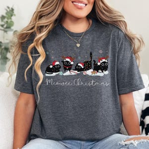 May include: Dark gray t-shirt featuring a festive design. The shirt displays four black cats wearing Santa hats and reindeer antlers, with the text "Meowee Christmas" in a whimsical font. Snowflakes and candy canes add to the holiday theme.
