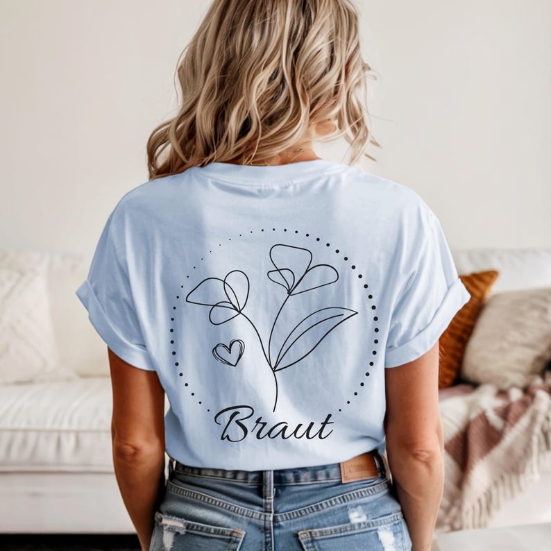 May include: Light blue t-shirt with a floral design and the word "Braut" in black script. The design features a line drawing of flowers, a heart, and a dotted circle. The shirt is made of soft material.