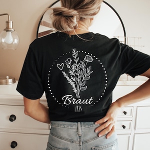 May include: Black t-shirt featuring a white floral bouquet design with the word "Braut" in script. The design is encircled by a dotted line and a heart. The shirt is a casual style.