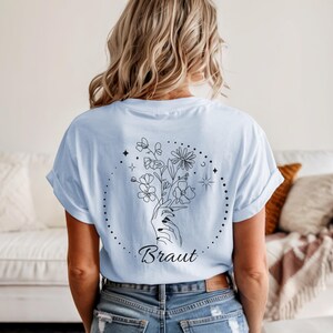Pu&ograve; includere: T-shirt azzurra con un design floreale nero e la scritta "Braut" stampata sul retro. Il design presenta una mano che tiene un bouquet di fiori all'interno di un cerchio tratteggiato, con stelle e altri elementi floreali.