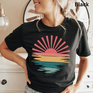May include: Black t-shirt featuring a retro sunset design. The graphic includes a pink sun with red rays, a yellow and orange horizon line, and blue water reflections. The word "Black" is printed at the top right.