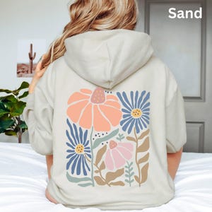 May include: A sand-colored hooded sweatshirt featuring a large graphic on the back. The design displays a collection of stylized flowers in shades of peach, blue, pink, and tan, with green stems and leaves. The word Sand is printed in the top right corner.
