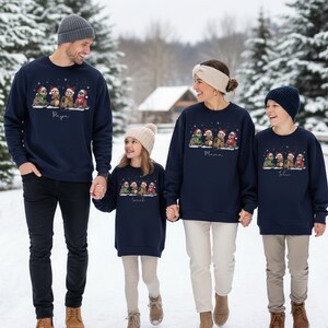 May include: Navy blue sweatshirts with a festive sloth design and personalized names. The sweatshirts feature a row of sloths wearing Santa hats and scarves. The family is holding hands in a snowy outdoor setting.