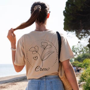May include: Tan t-shirt with a black floral design and the word "Crew" printed below. The design features two flowers and a heart within a dotted circle. The person is wearing blue jeans and a straw bag.