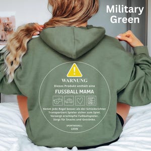 May include: Military green hoodie with a circular graphic. The graphic includes a warning symbol and text in German, "FUSSBALL MAMA," and icons. The text also mentions "SPORTMODELL: LEON."