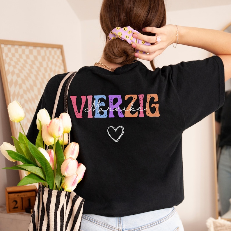 May include: Black t-shirt with the word "VIERZIG" in colorful block letters and the name "Marike" in cursive script. A white heart is below the text. The shirt is being worn by a person holding a striped tote bag with tulips.