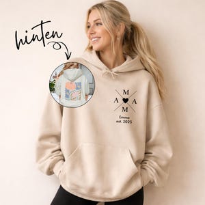 May include: A beige hoodie with a front pocket and a hood. The front features the letters "M A M" crossed with a heart in the center, and the text "Emma est. 2025". The back of the hoodie has a floral design.
