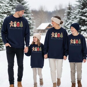 May include: A family of four wearing matching navy blue sweatshirts with a Christmas design. The design includes a gingerbread man, snowman, gift, and Christmas tree. The words "Merry Christmas" are printed below the design.