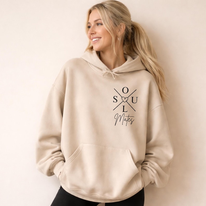 May include: A beige hooded sweatshirt with a front pocket and a drawstring hood. The sweatshirt displays the text "SOUL Mates" in a black, stylized font with a heart symbol. The garment is made of a soft material.