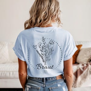 May include: Light blue t-shirt with a floral bouquet design and the word "Braut" in black script. The design includes a heart and a dotted circle. The shirt is a casual style.