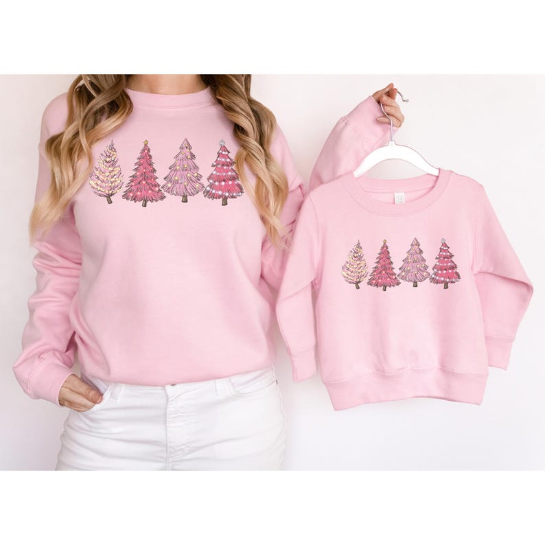 K&ouml;nnte beinhalten: Passende rosa Sweatshirts mit Weihnachtsb&auml;umen. Das Sweatshirt f&uuml;r Erwachsene zeigt f&uuml;nf B&auml;ume in Rosa- und Wei&szlig;t&ouml;nen. Das Sweatshirt f&uuml;r Kinder hat ein &auml;hnliches Design. Ein aufeinander abgestimmtes Kleidungsset.