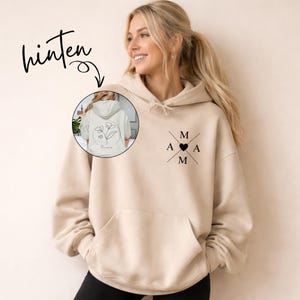 May include: A beige hoodie with a front pocket and a hood. The front features the letters "M A M" crossed with a heart in the middle. The back of the hoodie has a floral design inside a circle. The word "hinten" is written above the hoodie.