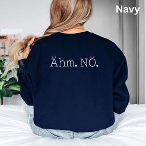 以下が含まれることがあります： 白で「&Auml;hm. N&ouml;.」とプリントされたネイビーブルーのスウェットシャツ。スウェットシャツは無地で、クルーネックです。テキストは衣服の背面に中央に配置されています。