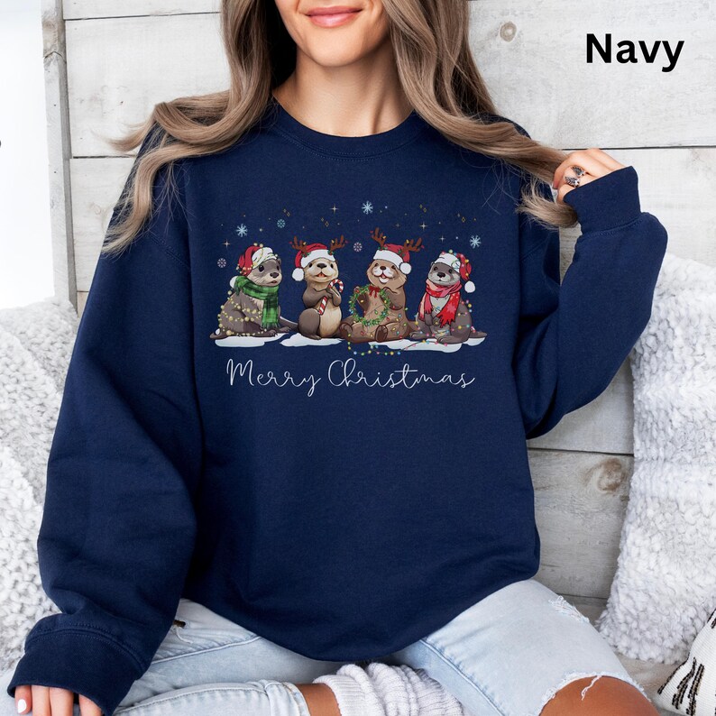 Pu&ograve; includere: Felpa blu navy con un design natalizio di lontre con cappelli e sciarpe festosi, decorate con luci a catena. La frase "Merry Christmas" &egrave; scritta in bianco sotto. La parola "Navy" &egrave; stampata in alto a destra.