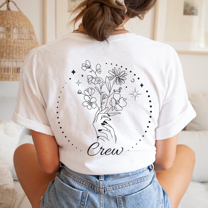 Pu&ograve; includere: T-shirt bianca con un disegno nero di una mano che tiene un mazzo di fiori all'interno di un cerchio tratteggiato. La parola "Crew" &egrave; scritta sotto. La maglietta &egrave; realizzata in un materiale morbido.