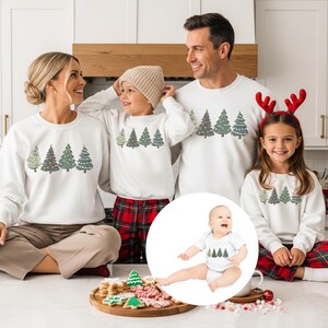 Puede incluir: Una familia de cuatro y un bebé visten sudaderas blancas con un diseño de árbol de Navidad. Las sudaderas tienen cuello redondo y mangas largas. El bebé lleva un body blanco con el mismo diseño de árbol. La familia sonríe y se mira.