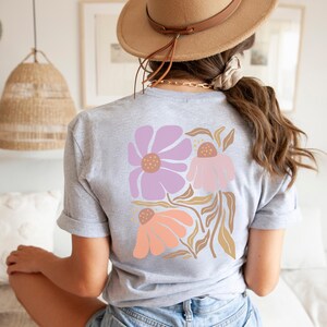 May include: A light gray short-sleeve t-shirt featuring a large graphic on the back. The design consists of three stylized flowers in shades of lavender, light pink, and peach, accompanied by tan-colored leaves and stems on a white background.