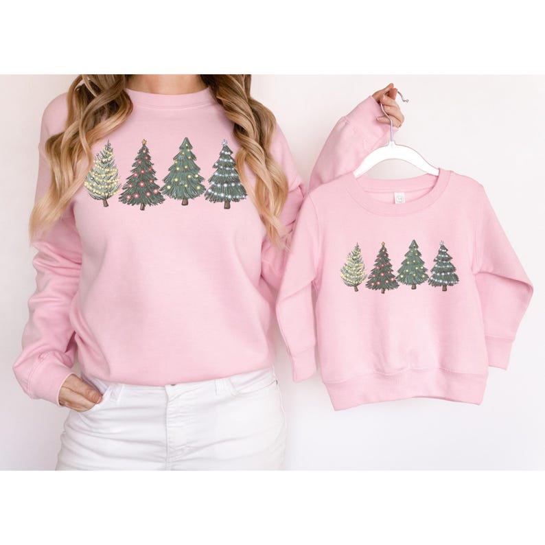 May include: Pink sweatshirts with a Christmas tree design. The adult sweatshirt features four different Christmas tree designs, while the child's sweatshirt has the same design. The sweatshirts are a light pink color.