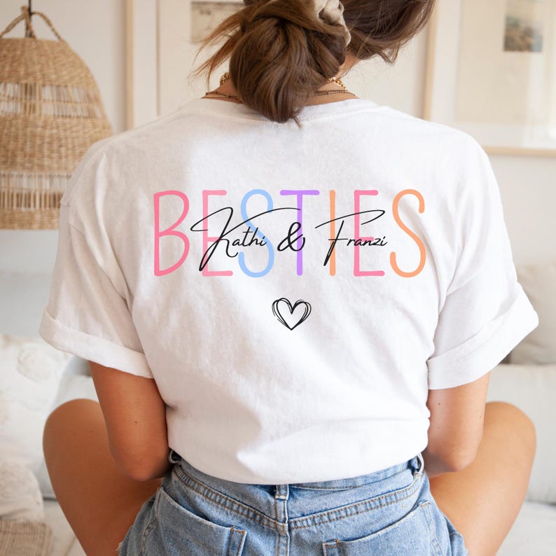 May include: White t-shirt featuring the word "BESTIES" in a rainbow of colours. The names "Kathi & Franzi" are written in black script, with a small heart underneath. The shirt is made of a soft fabric.