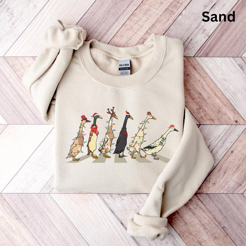 Peut inclure: Un sweat-shirt couleur sable avec un motif de sept canards portant des chapeaux de No&euml;l et des lumi&egrave;res. Les canards marchent sur un passage pour pi&eacute;tons.