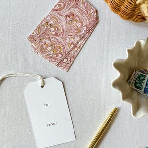 May include: A white gift tag with the words "TO:" and "FROM:" printed on it. The tag is tied with a white string and is sitting on a white surface. There is a stack of gift tags with a marbled pattern in shades of pink, brown, and gold in the background.