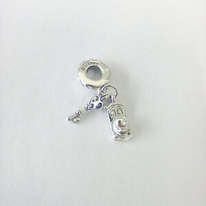 Könnte beinhalten: Ein silberner Charm-Anhänger mit einem Schlüssel und einem Türklopfer-Design. Der Charm hat oben eine Schlaufe zum Befestigen an einem Armband oder einer Halskette. Der Türklopfer hat ein Gesicht. Der Charm ist aus glänzendem Metall.