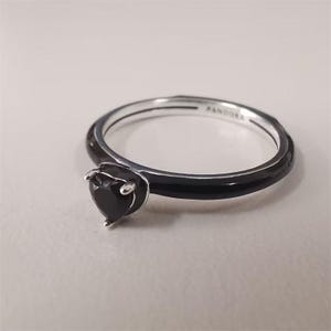 May include: A silver ring with a black enamel band and a heart-shaped black gemstone. The ring is marked with the brand name "PANDORA" on the inside of the band. The gemstone is set in a silver prong setting.