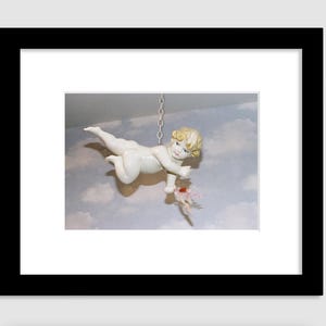 Puede incluir: Una figurita de querubín de porcelana blanca con cabello rubio, colgada de una cadena, vuela con los brazos extendidos, sosteniendo una flor rosa en su mano. La figurita está suspendida sobre un fondo de cielo nublado.