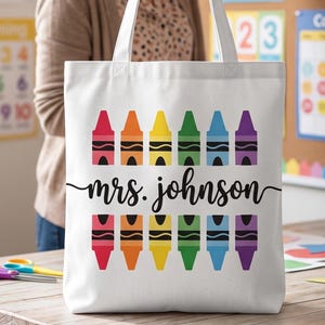 May include: A white canvas tote bag featuring two horizontal rows of colorful crayon illustrations in red, orange, yellow, green, blue, and purple. Centered between the rows is the black cursive text that reads mrs. johnson.