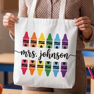May include: A white canvas tote bag featuring two rows of colorful crayon illustrations in red, orange, yellow, green, blue, and purple. The text "mrs. johnson" is printed in a black cursive script font centered between the two rows of crayons.
