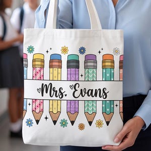 May include: A white canvas tote bag featuring a horizontal band across the center with the text Mrs. Evans in black script. The bag is decorated with a repeating pattern of colorful pencils in shades of yellow, purple, teal, and orange, accented with small flowers and stars.