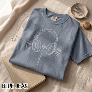 May include: A blue jean colored t-shirt featuring a white line art graphic of over-ear headphones with sound waves and the text "born for music" printed vertically below the headphones. The shirt has a crew neckline and short sleeves.