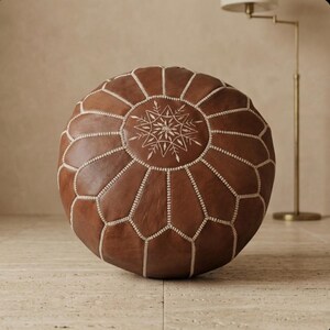 May include: Round brown leather ottoman with white stitching and a central starburst design. The ottoman is a decorative piece of furniture, perfect for a living room or bedroom. It is a comfortable and stylish seating option.