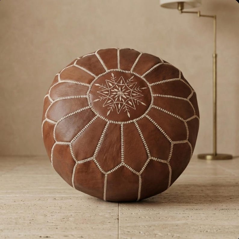 May include: Round brown leather ottoman with white stitching and a central starburst design. The ottoman is a decorative piece of furniture, perfect for a living room or bedroom. It is a comfortable and stylish seating option.