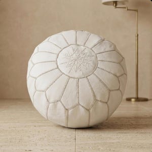 May include: A round, off-white leather pouf with a snowflake-like embroidered design in the center. The pouf is segmented with visible stitching and sits on a light-colored tiled floor. The design is a decorative accent for home decor.