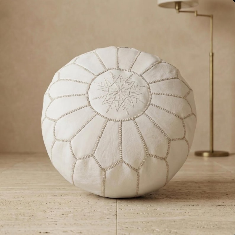 May include: A round, off-white leather pouf with a snowflake-like embroidered design in the center. The pouf is segmented with visible stitching and sits on a light-colored tiled floor. The design is a decorative accent for home decor.